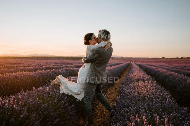 Vue latérale du marié soulevant mariée tout en se tenant dans le champ de lavande sur fond de ciel coucher de soleil le jour du mariage — Photo de stock