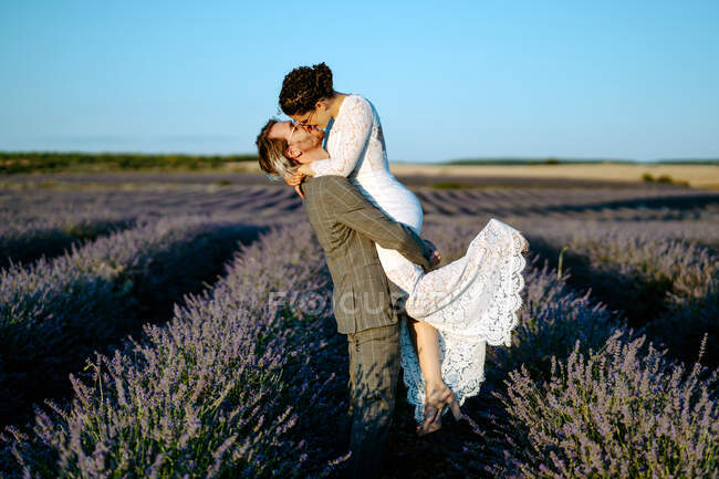 Vista lateral del novio levantando novia mientras está de pie en el campo de lavanda en el fondo del cielo azul claro en el día de la boda - foto de stock