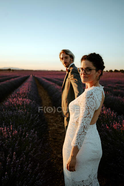 Наречений і наречений тримаються за руки і ходять у квітучому лавандовому полі, дивлячись на камеру. — стокове фото