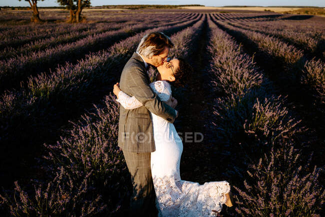 Visão lateral de alto ângulo de casal recém-casado romântico em pé cara a cara beijando no campo espaçoso contra o céu púrpura pôr do sol — Fotografia de Stock