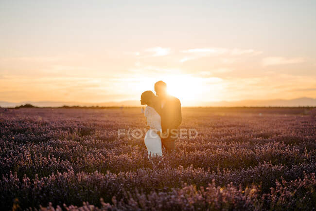 Seitenansicht des glücklichen Bräutigams umarmt Braut steht in Lavendelfeld vor dem Hintergrund des Sonnenuntergangs Himmel am Hochzeitstag — Stockfoto
