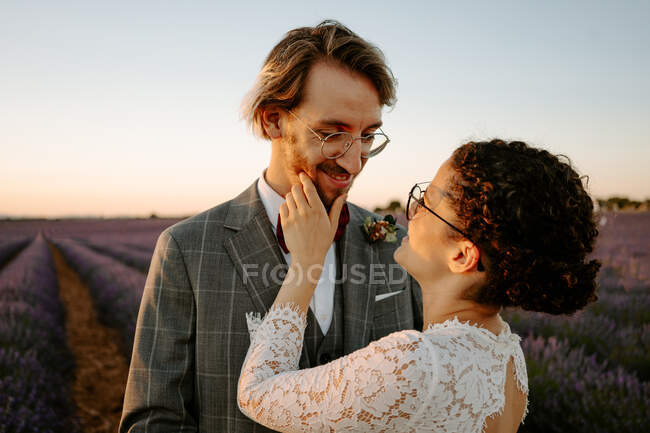 Zufriedene Braut im weißen Kleid berührt Gesicht des lächelnden Bräutigams im Anzug, während sie im Lavendelfeld bei Sonnenuntergang steht und in die Kamera schaut — Stockfoto