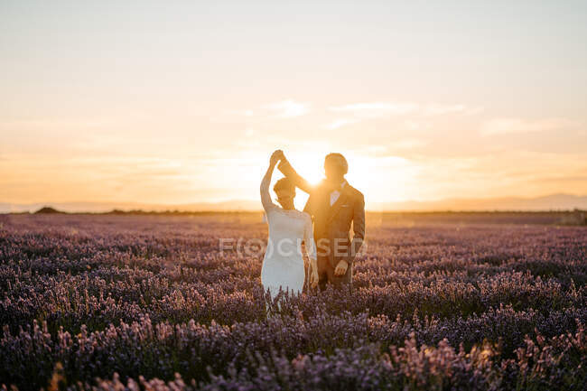 Щаслива безлика молода пара в стильних весільних сукнях танцює і тримає руки м'яко на просторому квітучому лавандовому полі на мальовничому заході сонця — стокове фото