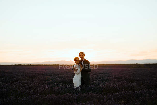 Боковой вид романтической молодоженов, стоящих лицом к лицу на просторном поле против фиолетового неба заката, смотрящих в камеру — стоковое фото