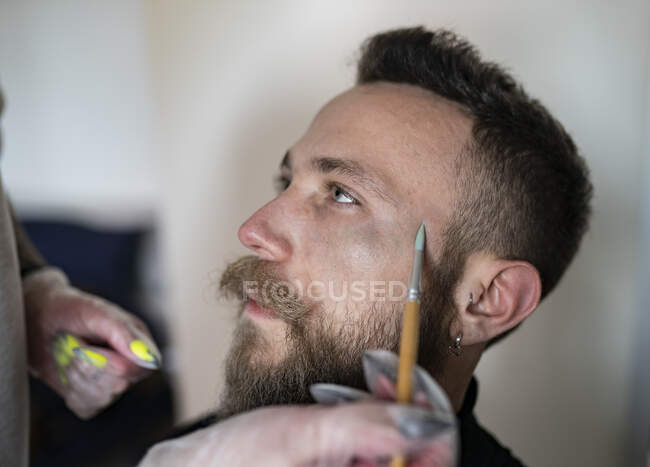 Крупним планом невпізнаваний візажист, який кладе макіяж на обличчя хіпстерського чоловіка — стокове фото