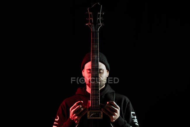 Tranquil male rock musician standing with electric guitar on black background in studio with red light — Stock Photo