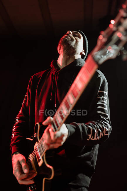 Low angle of rock guitarist playing electric guitar while performing in dark studio with red light — Stock Photo