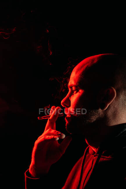 Vista laterale del rocker maschio con testa calva fumante ed espirante fume in studio scuro con luce rossa al neon su sfondo nero — Foto stock