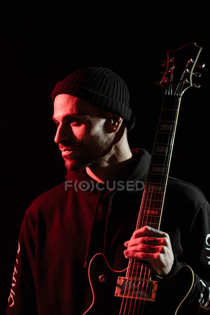 Tranquil male rock musician standing with electric guitar on black background in studio with red light — Stock Photo