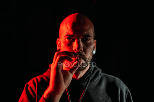Серйозний чоловічий рокер палить сигарету і дивиться на камеру на чорному тлі в студії з червоним освітленням — стокове фото