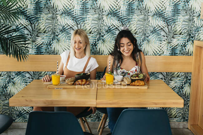 Junge zufriedene Freundinnen sitzen am Holztisch mit leckeren Croissants und Getränken in der Cafeteria — Stockfoto