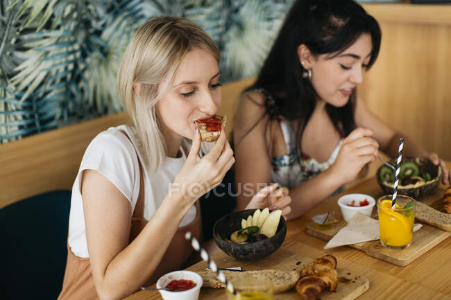 Giovani contenuti migliori amiche sedute a tavola di legno con gustosi croissant e bevande in mensa — Foto stock