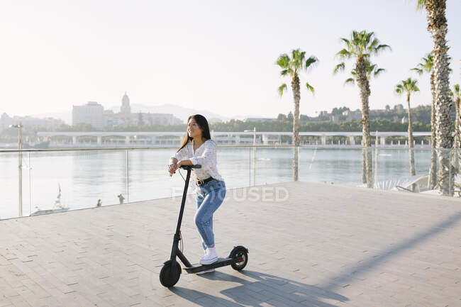Joven mujer asiática alegre en ropa casual montando scooter eléctrico contra el río mientras mira hacia adelante en la ciudad - foto de stock