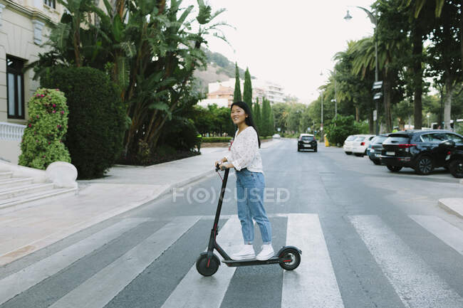 Коли дивишся на фотоапарат, то бачиш, як молода азіатка їде на електричному скутері по міській дорозі проти зелених дерев. — стокове фото