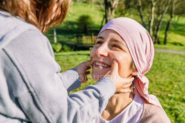 Unrecognizable daughter drawing smile with fingers on mother's face with cancer wearing pink head scarf standing on green park looking at each other — Stock Photo
