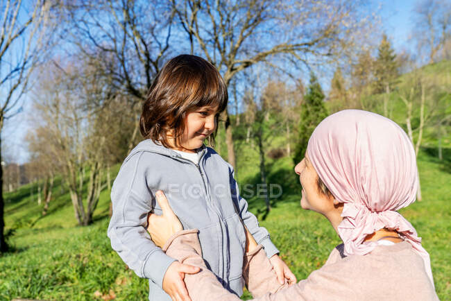Vista lateral de madre feliz con cáncer con pañuelo rosa en la cabeza sosteniendo a la pequeña hija de pie en el parque verde mirándose el uno al otro - foto de stock