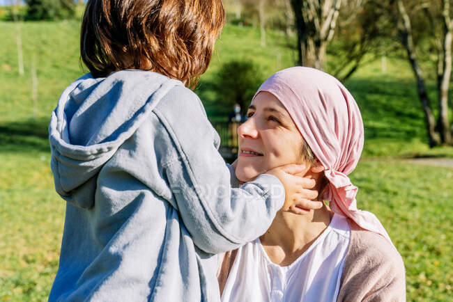 Unerkennbare Tochter zeichnet Lächeln mit den Fingern auf dem Gesicht der krebskranken Mutter, die ein rosafarbenes Kopftuch trägt und im grünen Park steht und einander anschaut — Stockfoto