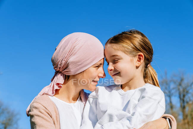 Счастливая мать с раком в розовом шарфе обнимает маленькую дочь в зеленом парке, глядя друг на друга. — стоковое фото