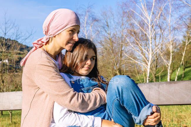 Glückliche krebskranke Mutter mit rosa Kopftuch umarmt kleine Tochter, die auf einer Bank im grünen Park sitzt und wegschaut — Stockfoto