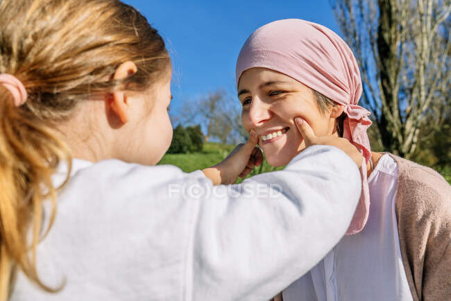 Невпізнавана дочка малює посмішку з пальцями на обличчі матері з раком в рожевому шарфі голови, що стоїть на зеленому парку, дивлячись один на одного — стокове фото