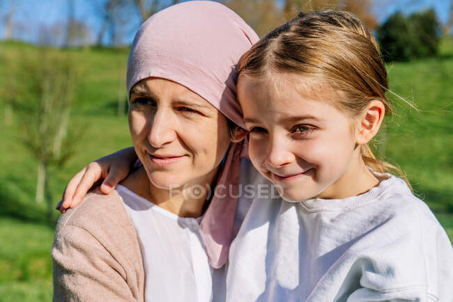 Mãe feliz com câncer vestindo cachecol cabeça rosa abraçando pequena filha no parque verde olhando para longe — Fotografia de Stock