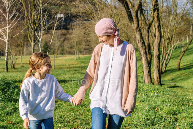 Madre feliz con cáncer con pañuelo rosa en la cabeza tomados de la mano con su hija pequeña mientras caminan en el parque verde mirándose entre sí - foto de stock