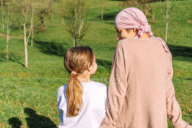 Вид ззаду ніжна спокійна мати з раком в рожевому шарфі голови, що говорить з маленькою донькою, сидить на лавці на зеленому парку — стокове фото