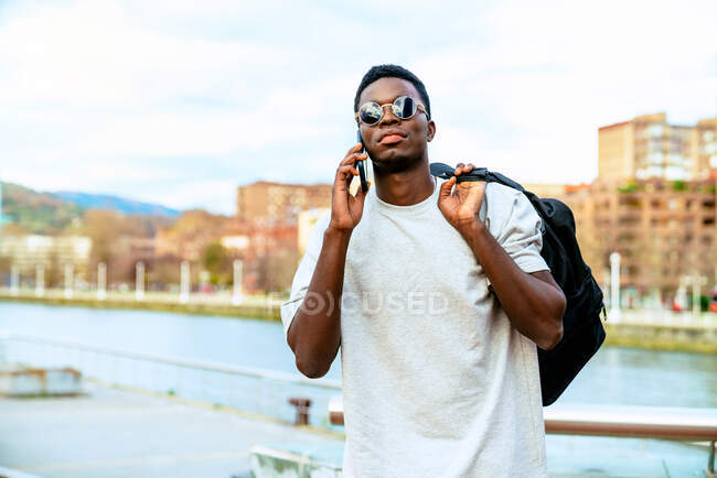 Jovem afro-americano macho com mochila na moda óculos de sol falando no celular contra o rio da cidade sob céu nublado — Fotografia de Stock
