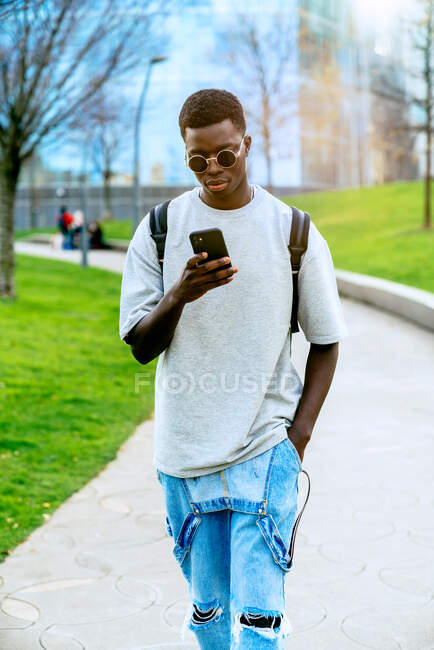 Joven y elegante hombre afroamericano con la mano en el bolsillo navegando por Internet en el teléfono celular mientras camina por el camino en la ciudad - foto de stock