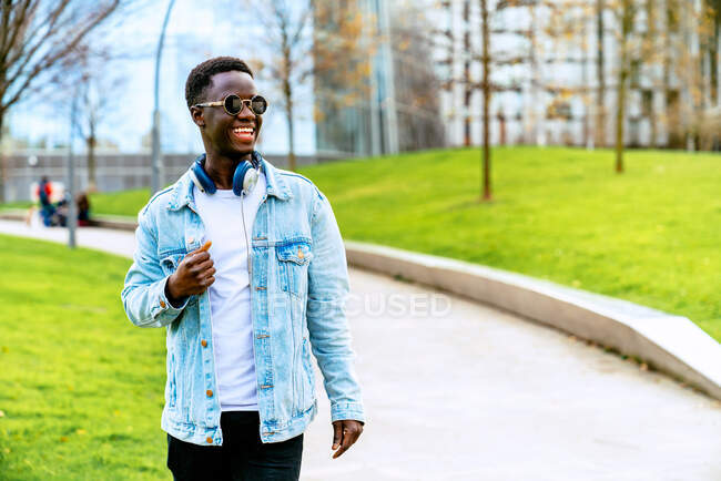 Contenido joven Hombre afroamericano en chaqueta de mezclilla con auriculares en el camino entre el césped en la ciudad - foto de stock