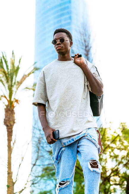 Bajo ángulo de joven masculino afroamericano en overoles de mezclilla con smartphone y mochila en la ciudad - foto de stock
