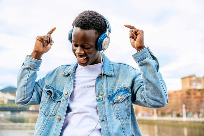 Young content African American male in headset listening to song while dancing with raised arms under cloudy sky in town — Stock Photo