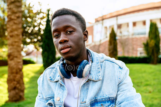 Young black male in denim jacket with headset looking at camera on green meadow in city — Stock Photo