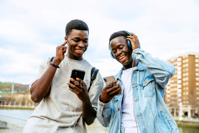 Socios étnicos de contenido joven con teléfonos celulares escuchando canciones de auriculares y auriculares en el terraplén urbano bajo el cielo nublado - foto de stock