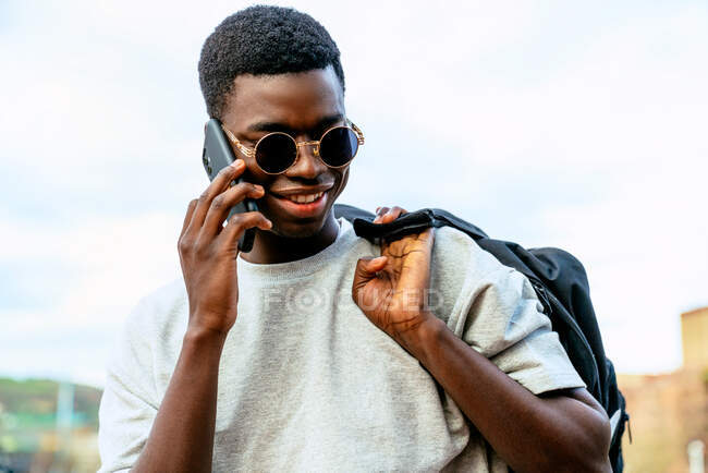 Junger Afroamerikaner mit Rucksack und modischer Sonnenbrille im Gespräch auf einem Handy unter wolkenverhangenem Himmel. — Stockfoto