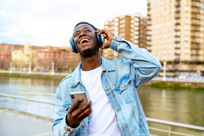Joven hombre negro positivo con teléfono celular escuchando la canción de los auriculares mientras mira hacia arriba en terraplén urbano - foto de stock