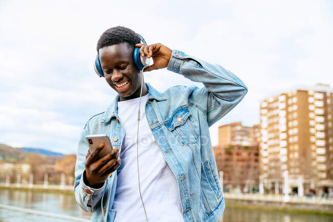 Jovem macho negro positivo com celular ouvindo música de fones de ouvido enquanto olha para cima no aterro urbano — Fotografia de Stock