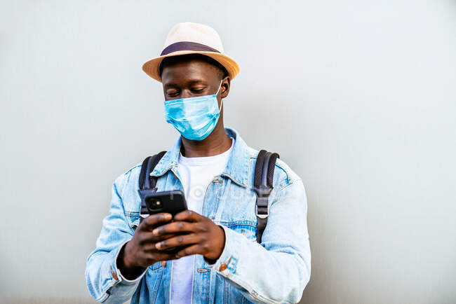 Anonymous focused ethnic male in hat and face mask text messaging on cellphone on light background — Stock Photo