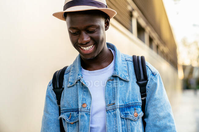 Joven hombre negro alegre en chaqueta de mezclilla mirando hacia otro lado con sonrisa dentada a la luz del día - foto de stock