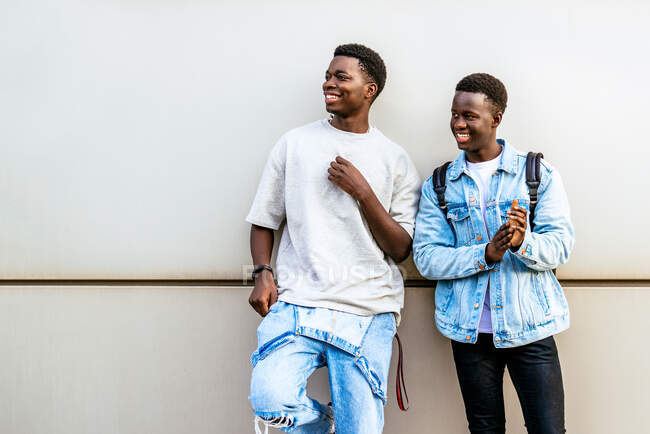 Joyeux jeunes amis afro-américains en vêtements décontractés regardant sur le côté sur un mur de la ville. — Photo de stock