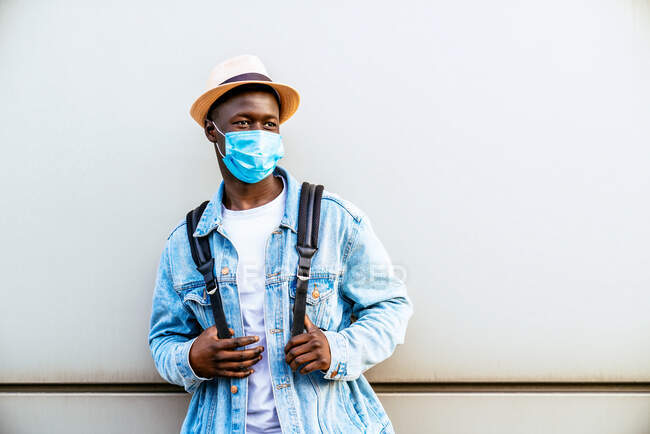 Juventud afroamericana anónima con una máscara estéril y ropa casual mirando hacia un lado en una pared de la ciudad durante la pandemia de COVID 19. - foto de stock