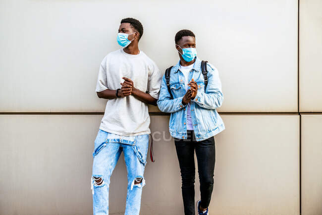 Anonymous young ethnic male partners in face masks and trendy outfit looking away near wall in daytime — Stock Photo