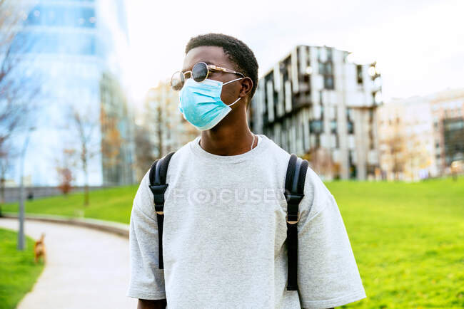 Hombre afroamericano irreconocible con máscara estéril y gafas de sol mirando hacia otro lado en la pasarela entre el césped brillante de la ciudad durante la pandemia de COVID 19 - foto de stock