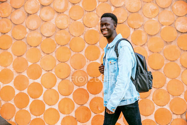 Jovem negro com jaqueta de ganga e mochila olhando para a câmera enquanto caminha — Fotografia de Stock