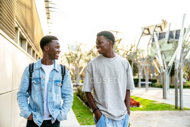 Jeunes amis afro-américains heureux en vêtements décontractés avec les mains dans les poches se regardant les uns les autres sur la passerelle en ville — Photo de stock