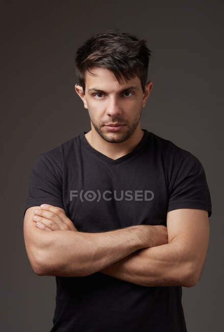 Homme en chemise noire avec bras croisés regardant la caméra sur fond gris en studio — Photo de stock