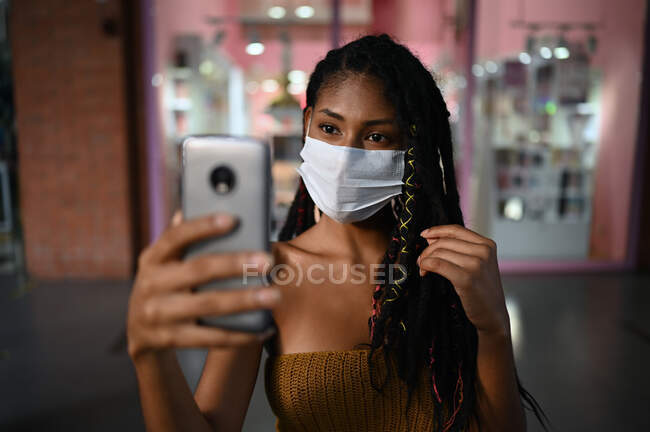 Portrait of attractive young afro latin woman wearing a facemask takes selfie with smartphone in a commercial mall, Colombia — Stock Photo
