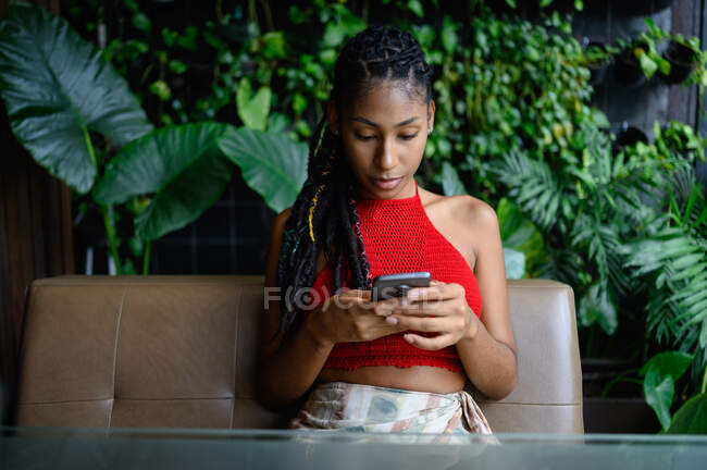 Retrato de mujer latina afro joven atractiva con rastas en un top rojo del ganchillo usando smartphone en mesa de restaurante, Colombia - foto de stock