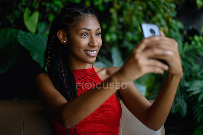 Retrato de atraente jovem afro mulher latina com dreadlocks em um top vermelho de crochê usando smartphone na mesa de restaurante, Colômbia — Fotografia de Stock