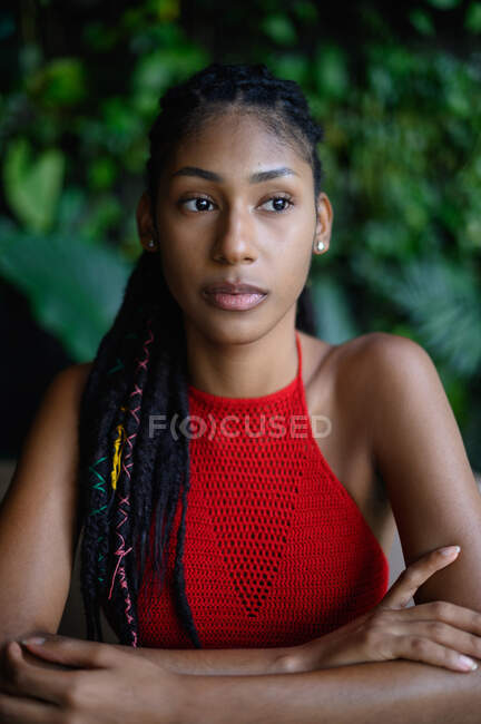 Retrato de mujer latina afro contemplativa joven con rastas en un top rojo del ganchillo sentado en la mesa, Colombia - foto de stock